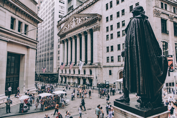 Scena di strada affollata nel Financial District di New York, con una grande statua in primo piano e l'edificio della Borsa di New York con bandiere americane sullo sfondo, circondato da persone e venditori.