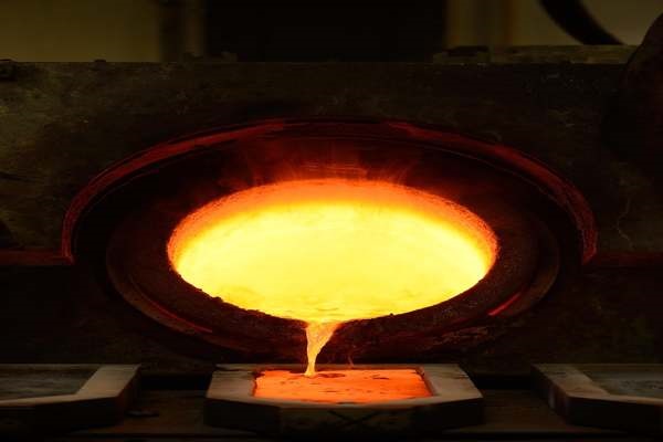 Il metallo fuso viene versato da una fornace incandescente negli stampi sottostanti, con un'intensa luce arancione e gialla che illumina la scena.