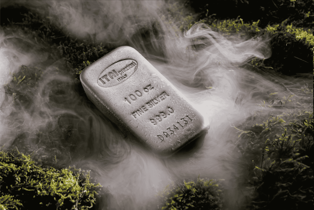 A 100 oz silver bar lies on green moss, surrounded by swirling, misty fog. The bar is stamped with purity and weight details and appears metallic and shiny amidst the natural, moody setting.