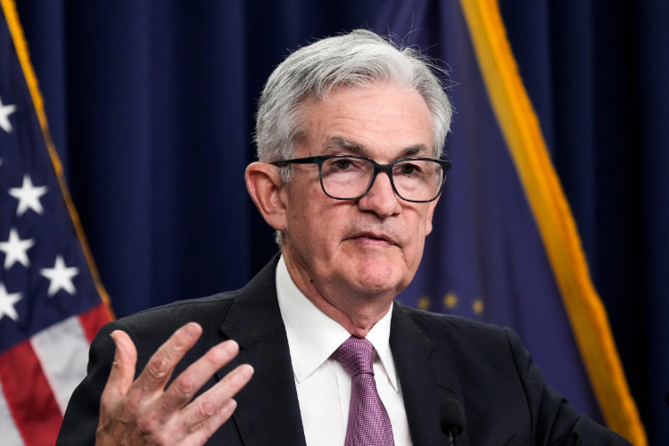 Un uomo in giacca e cravatta, con capelli grigi e occhiali, parla da un podio gesticolando con una mano; sullo sfondo sono visibili una bandiera americana e una tenda blu scuro.