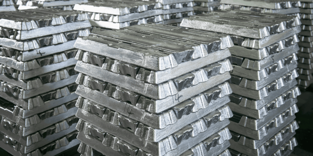 Pile di lingotti di metallo argentato lucido sono disposte in pile ordinate, creando un motivo geometrico. I lingotti hanno una consistenza ruvida e una lucentezza metallica che riflette la luce dall'alto.