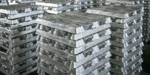 Pile di lingotti di metallo argentato lucido sono disposte in pile ordinate, creando un motivo geometrico. I lingotti hanno una consistenza ruvida e una lucentezza metallica che riflette la luce dall'alto.