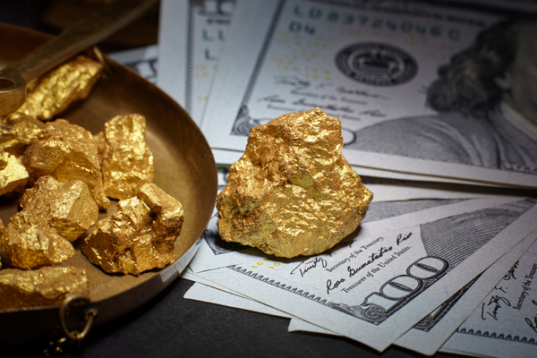 Diverse pepite d'oro sono appoggiate su una bilancia di bronzo e accanto a banconote da 100 dollari sparse, a simboleggiare ricchezza e valore.