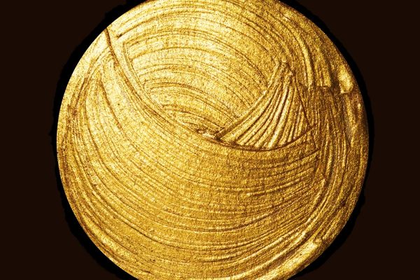 Un vortice circolare di vernice dorata lucida e metallica su uno sfondo nero pieno, con pennellate curve e strutturate che creano un motivo astratto e scintillante.