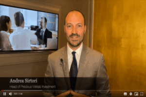 Un uomo in giacca e cravatta si trova al chiuso e parla alla telecamera con un microfono agganciato alla giacca. Dietro di lui, uno schermo mostra persone in riunione d'affari. Il testo recita: Andrea Sirtori, Responsabile Investimenti Metalli Preziosi.