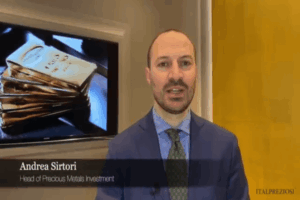 Un uomo in giacca e cravatta si trova di fronte a una parete gialla e a un televisore che mostra lingotti d'oro. Il testo dell'immagine recita: Andrea Sirtori, Responsabile Investimenti Metalli Preziosi. Il logo ITALPREZIOSI appare nell'angolo in basso a destra.