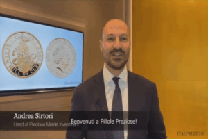 Un uomo in giacca e cravatta sta in casa e sorride alla telecamera. Dietro di lui, uno schermo mostra due monete d'oro. Il testo dell'immagine recita: Andrea Sirtori, Head of Precious Metals Investment e Benvenuti a Pillole Preziose.