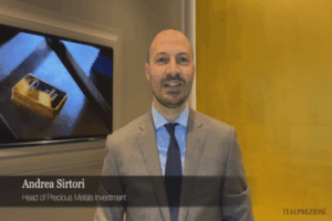 Un uomo in giacca e cravatta sta in casa e sorride alla telecamera. Dietro di lui c'è un televisore che mostra un lingotto d'oro e un muro giallo. Il testo dell'immagine recita: Andrea Sirtori, Responsabile Investimenti Metalli Preziosi, ITALPREZIOSI.