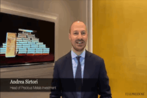 Un uomo in giacca e cravatta è in piedi, sorridente, accanto a uno schermo che mostra lingotti d'oro. Il testo sullo schermo recita: Andrea Sirtori, Responsabile Investimenti Metalli Preziosi. Il logo di Italpreziosi appare in basso a destra.