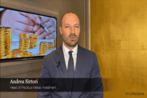 Un uomo in giacca e cravatta si trova in un ufficio. Dietro di lui, uno schermo mostra monete d'oro impilate e mani che tengono una penna. Un riquadro di testo recita Andrea Sirtori, Responsabile Investimenti Metalli Preziosi, ITALPREZIOSI.