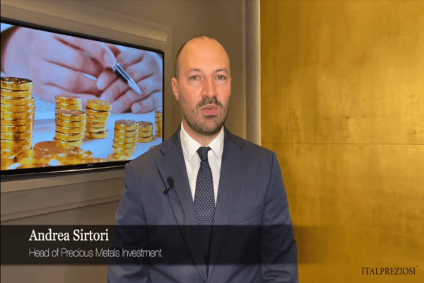 Un uomo in giacca e cravatta si trova in un ufficio. Dietro di lui, uno schermo mostra monete d'oro impilate e mani che tengono una penna. Un riquadro di testo recita Andrea Sirtori, Responsabile Investimenti Metalli Preziosi, ITALPREZIOSI.