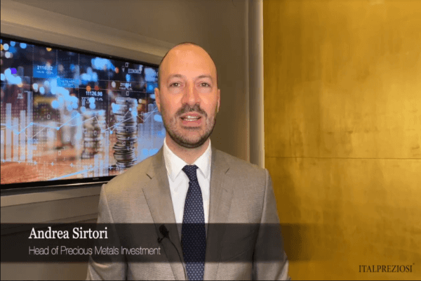 Un uomo in giacca e cravatta grigia si trova in un ufficio con alle spalle uno schermo che visualizza grafici finanziari e pile di monete. Il testo in sovrimpressione recita Andrea Sirtori, Responsabile Investimenti Metalli Preziosi, ITALPREZIOSI.