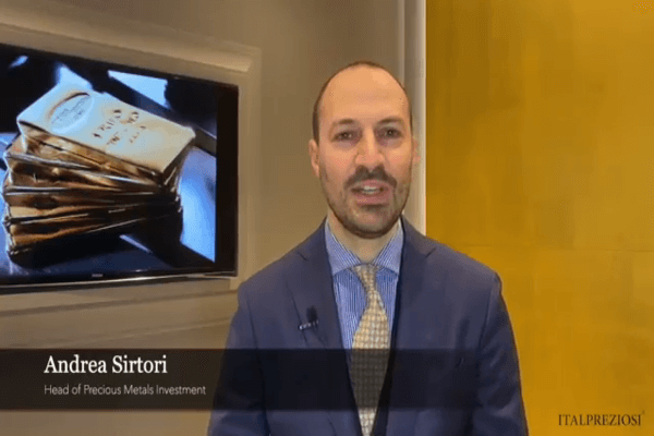 Un uomo in giacca e cravatta blu è in piedi al chiuso; dietro di lui un televisore mostra lingotti d'oro impilati. Il testo sullo schermo recita Andrea Sirtori, responsabile degli investimenti in metalli preziosi, con ITALPREZIOSI nell'angolo.