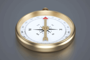 Una bussola con cornice dorata, quadrante bianco, segni di direzione neri e ago con punta rossa che punta a nord, appoggiata su una superficie grigia.