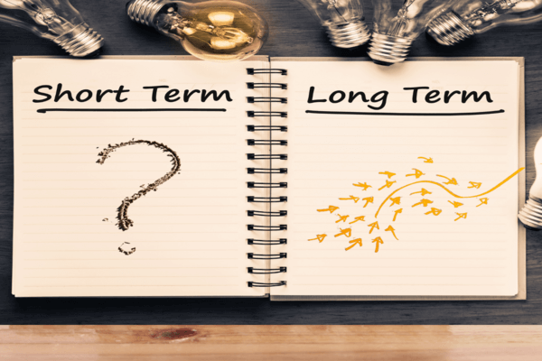 Un quaderno aperto è suddiviso in Breve termine con un punto interrogativo e Lungo termine con frecce che si muovono verso l'alto e verso l'esterno; le lampadine sono sparse intorno al quaderno su una superficie scura.