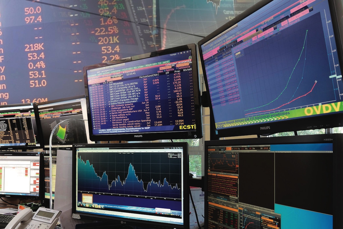 Varios monitores de ordenador muestran cuadros financieros, gráficos y datos bursátiles en una sala de negociación, lo que indica un análisis y seguimiento activos del mercado.