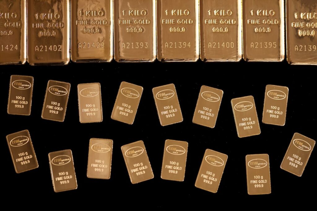 En la parte superior se alinean varios lingotes de oro de gran tamaño con la etiqueta 1 KILO DE ORO FINO, y debajo se disponen en filas lingotes de oro más pequeños con la etiqueta 100 g DE ORO FINO, todo sobre fondo negro.