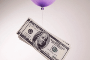 Un palloncino viola è legato a un fascio di banconote da cento dollari statunitensi, dando l'impressione che il denaro venga sollevato in aria. Lo sfondo è semplice e di colore chiaro.