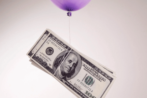 Un palloncino viola è legato a un fascio di banconote da cento dollari statunitensi, dando l'impressione che il denaro venga sollevato in aria. Lo sfondo è semplice e di colore chiaro.