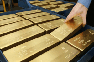 La mano di una persona che raccoglie uno dei tanti lingotti d'oro di grandi dimensioni ordinatamente impilati in un contenitore metallico blu; i lingotti d'oro hanno iscrizioni e numeri di serie incisi sulla superficie.