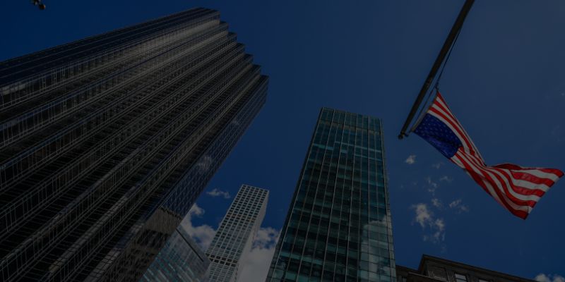 Grattacieli visti dal basso contro un cielo azzurro, con una bandiera americana appesa sul lato destro dell'immagine.
