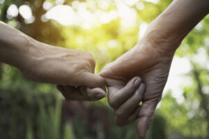 Two people gently holding hands outdoors, with sunlight shining through green trees in the background, symbolizing connection, support, or love.