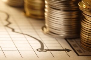Primo piano di monete impilate su un grafico finanziario con una linea di tendenza al ribasso e una freccia, che simboleggia il declino economico o la perdita finanziaria. L'illuminazione calda mette in risalto le monete metalliche e il grafico cartaceo.