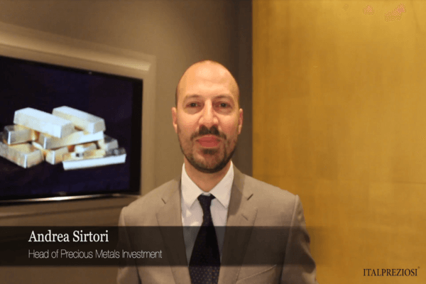 Un uomo in giacca e cravatta sta in casa e sorride alla telecamera. Dietro di lui c'è un'immagine incorniciata di lingotti d'oro. Il testo recita: Andrea Sirtori, Responsabile Investimenti Metalli Preziosi e ITALPREZIOSI nell'angolo.