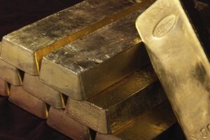 Diversi lingotti d'oro lucidi sono impilati l'uno sull'altro su una superficie scura, con i lingotti che riflettono la luce e mostrano alcuni segni incisi.