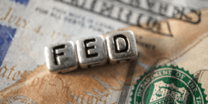 Tre dadi in metallo compongono la scritta FED, che rappresenta la Federal Reserve statunitense. Sullo sfondo una banconota di dollari statunitensi.