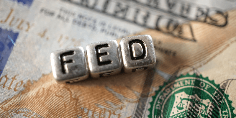 Tre dadi in metallo compongono la scritta FED, che rappresenta la Federal Reserve statunitense. Sullo sfondo una banconota di dollari statunitensi.